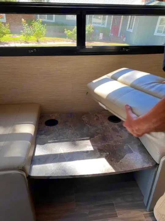 RV rental dinette: A person adjusts light-colored cushions on a table with cup holders inside the vehicle.. Thor Axis 2023