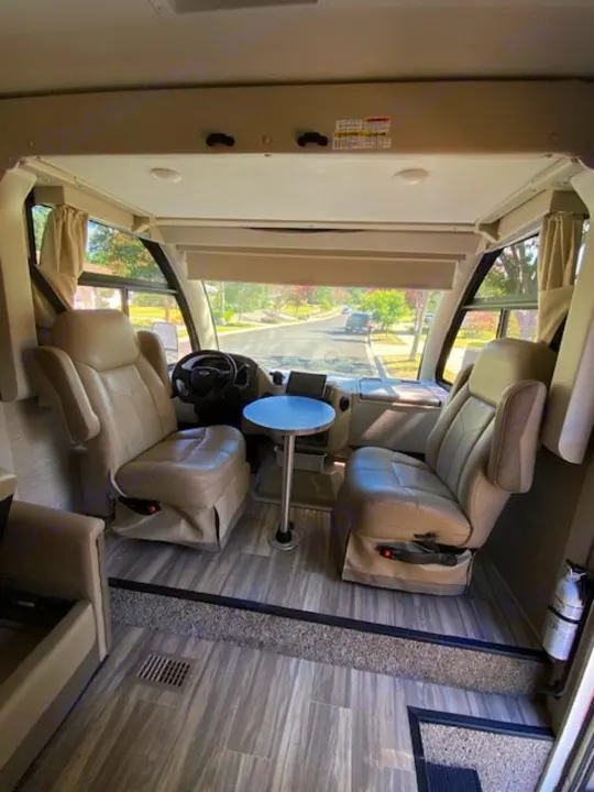 Front view of an RV rental showing its spacious cockpit: two captain's chairs, small table, and road ahead through the windshield.. Thor Axis 2023
