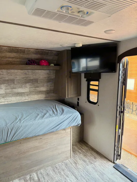 RV rental interior: bed with grey sheet, TV, AC unit, and storage shelf with helmets. Ready for your adventure.. Keystone RV Springdale 2022