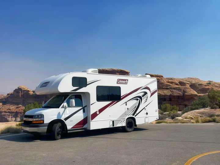 White Coleman RV rental on a paved lot, with rugged desert rock formations under a clear blue sky. Ideal for adventure travel.. Thor Motor Coach Coleman 2021