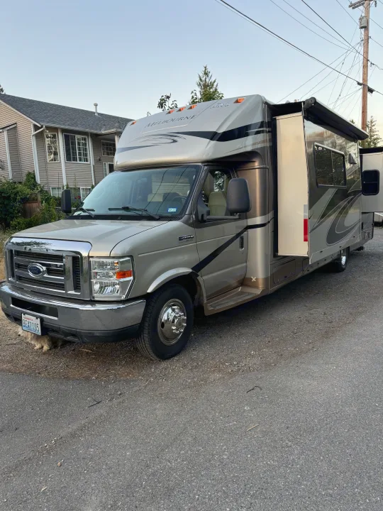 A light-colored RV rental is parked on a gravel lot near a house, with its slide-out extended and a small dog under the front.. Jayco Melbourne 2012