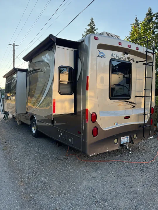 A beige and brown Jayco Melbourne RV rental, its side extensions open, is parked on gravel. A small dog is visible beneath it.. Jayco Melbourne 2012