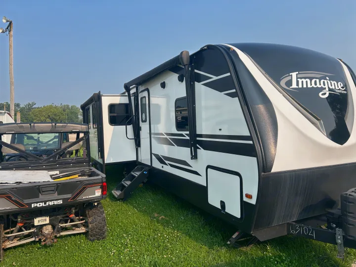 RV rental: A white and black Imagine travel trailer with an extended awning, parked on grass next to a Polaris UTV.. Grand Design Imagine 2021