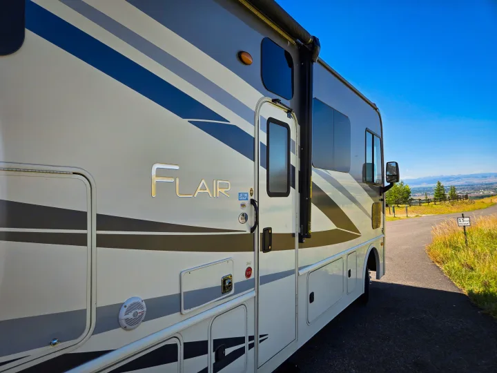 This Flair motorhome RV rental, featuring blue and gray stripes, is parked on a road with scenic mountain views.. Flair-Fleetwood Flair-Fleetwood Motorhome 2019