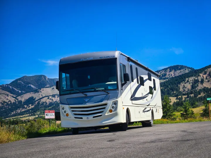 A cream and grey Fleetwood Flair RV rental on a mountain road under a bright blue sky, with pine-covered mountains in the background.. Flair-Fleetwood Flair-Fleetwood Motorhome 2019