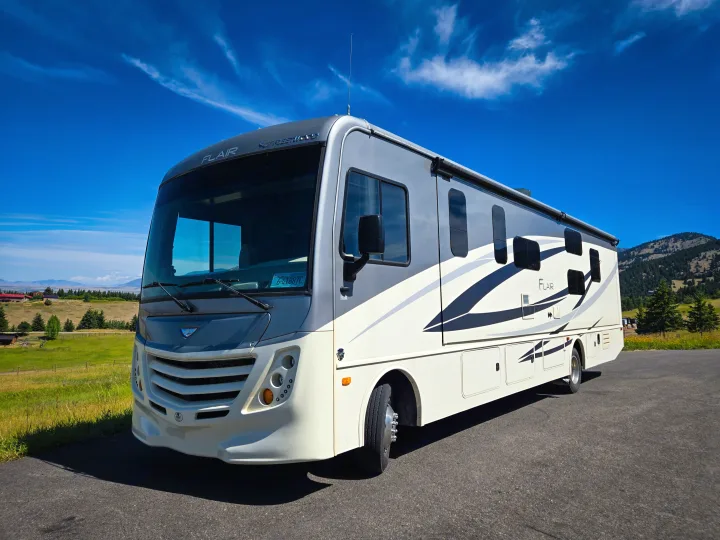 A modern Flair RV rental, cream and gray, is parked on asphalt with green fields, distant mountains, and a clear blue sky.. Flair-Fleetwood Flair-Fleetwood Motorhome 2019