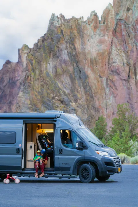 A family enjoys their RV rental with a child and balance bike, relaxing in their camper van door by stunning mountains.. Coachmen PIXEL 2026