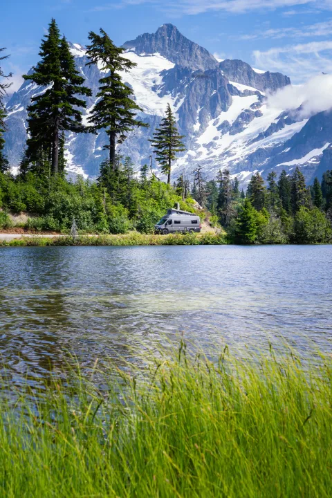 A gray RV rental parked by a peaceful lake, surrounded by a lush forest and majestic snow-capped mountains.. Coachmen PIXEL 2026