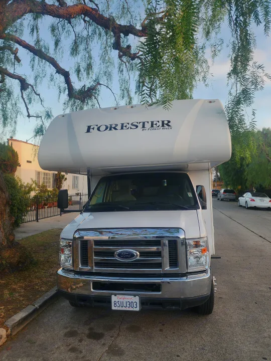 A white Ford Forester Class C RV (camper van) is parked on a quiet, tree-lined street. An excellent rv rental option.. Forest River Forester 2018