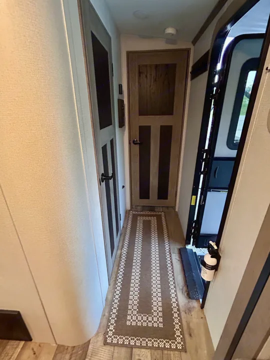RV rental interior: A narrow hallway with two doors, a runner rug, and an open entry door with steps and a fire extinguisher.. Keystone RV Outback 2021