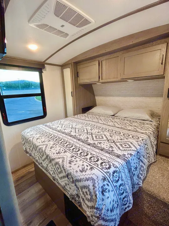 Cozy RV rental bedroom with patterned bedspread, overhead cabinets, and a scenic window view.. Keystone RV Outback 2021