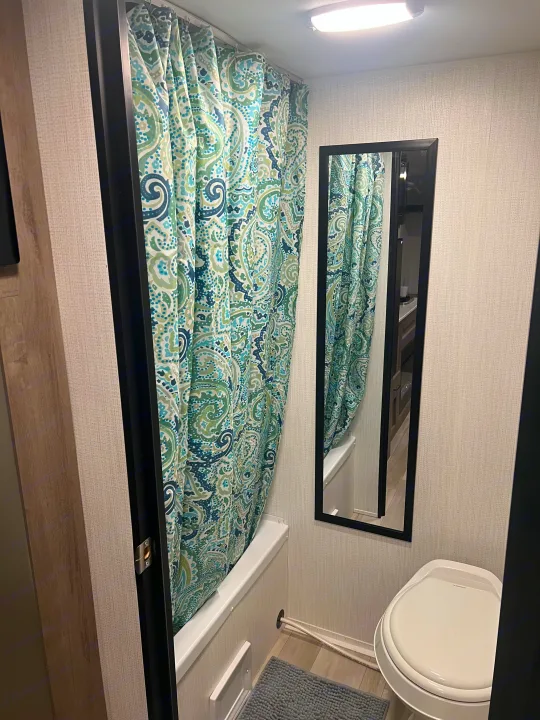 Inside an RV rental, a compact bathroom features a colorful paisley shower curtain, white toilet, and black-framed mirror.. Heartland Prowler Lynx 17BHX 2025