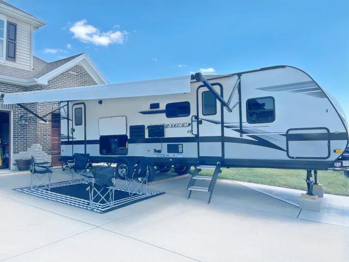 Modern white & gray travel trailer RV rental set up on a driveway with extended awning, rug, and chairs.. Jayco Jay Feather 2022