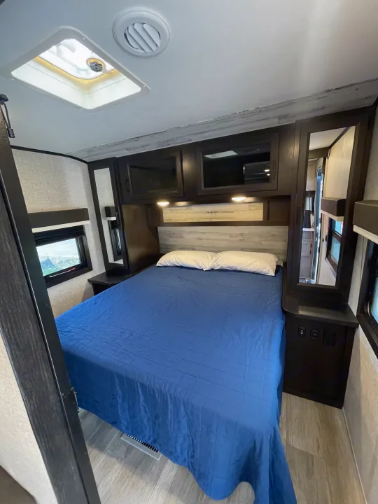 Cozy RV rental bedroom with a blue-comforter bed, white pillows, overhead storage, and light-filled windows.. Jayco Jay Feather 2022