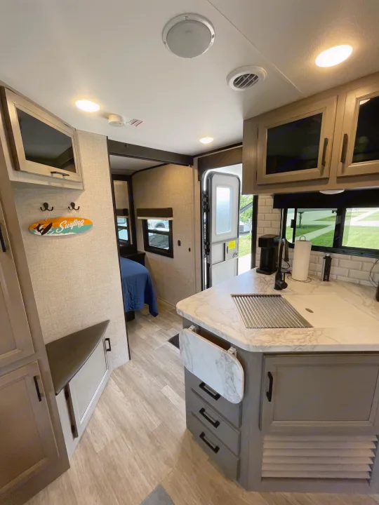 Bright RV rental interior: kitchen with sink, extendable counter, storage, and surfing decor; bedroom beyond.. Jayco Jay Feather 2022
