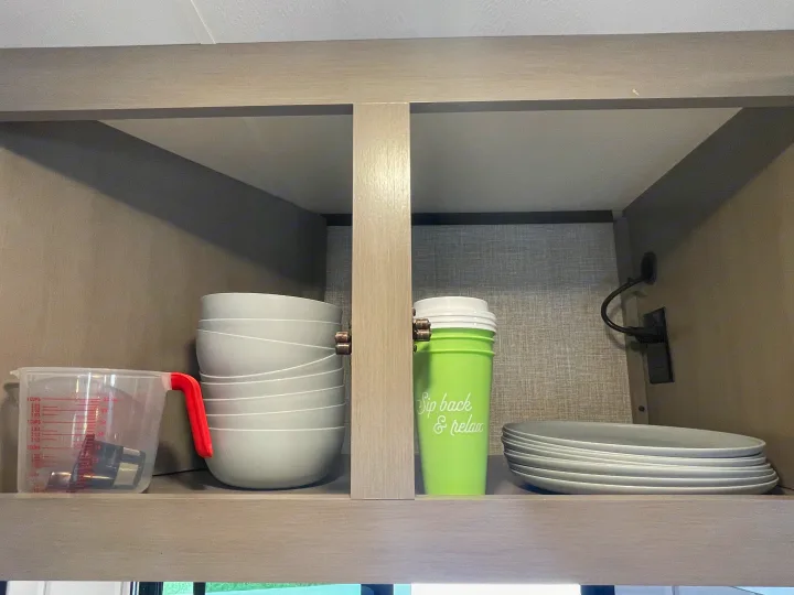 RV rental kitchen cabinet showing organized dishes: a measuring cup, stacked bowls, a travel mug, and stacked plates.. Jayco Jay Feather 2022