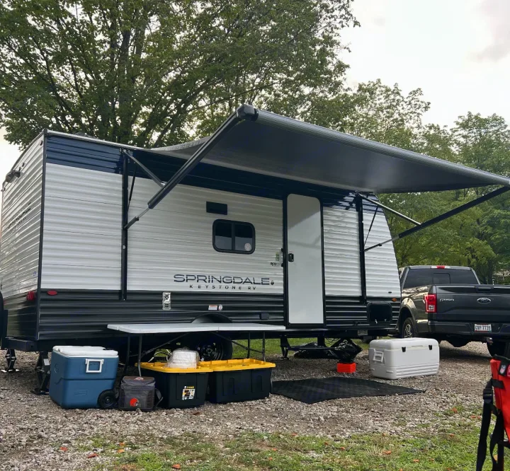 This **RV rental** is set up for camping, showcasing a white travel trailer with an extended awning, coolers, and a truck parked nearby.. Keystone RV Springdale Mini 2025