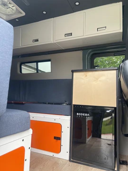 Inside this RV rental, find a compact interior with light wood flooring, white and orange cabinets, blue cushioned seating, and an integrated cooler.. Mercedes Sprinter 2021