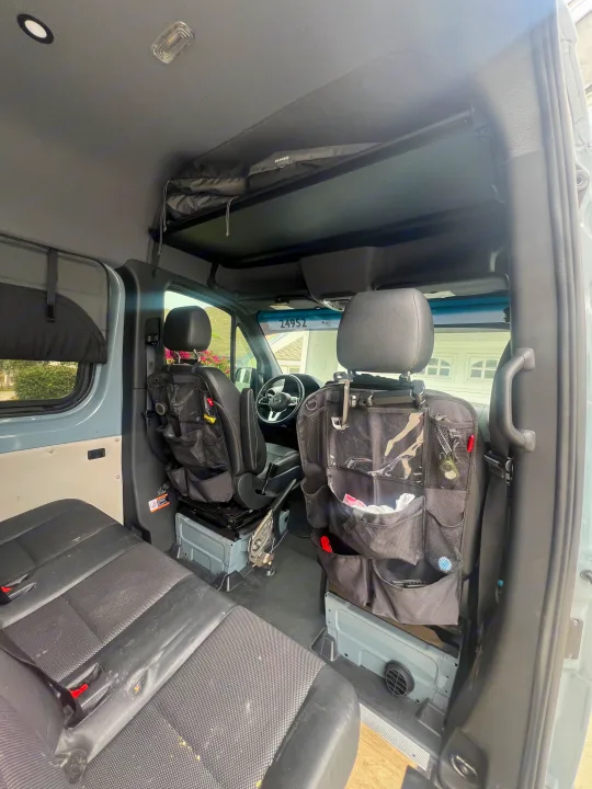 An RV rental's spacious interior from the rear. Front seats, rear bench, and overhead storage are visible.. Mercedes Sprinter 2021