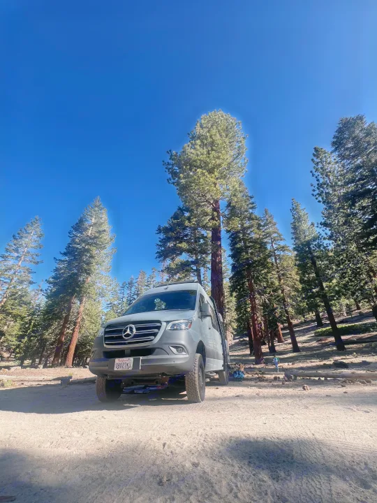 A grey Mercedes Sprinter rv rental van is parked on a dirt road amidst towering pine trees under a clear blue sky, ideal for outdoor trips.. Mercedes Sprinter 2021