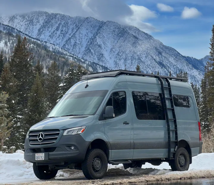 Sleek grey RV rental: a Mercedes Sprinter van parked on a snow-covered road with majestic, pine-dotted mountains. Perfect for winter adventures.. Mercedes Sprinter 2021
