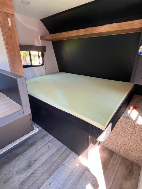 Interior of an RV rental showing a bed with a foam mattress topper, storage shelf, and wood flooring.. Keystone RV Springdale 2022