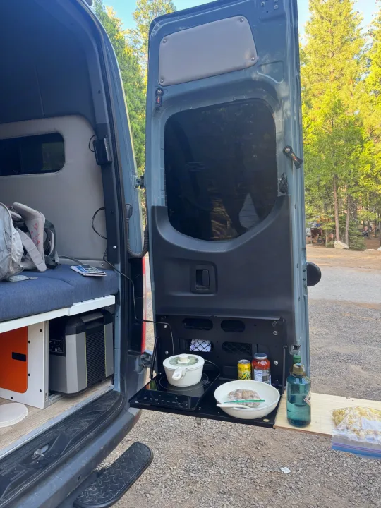 RV rental: A camper van's open rear reveals an outdoor kitchen with a cooktop, food, and power station, set in a peaceful campsite.. Mercedes Sprinter 2021