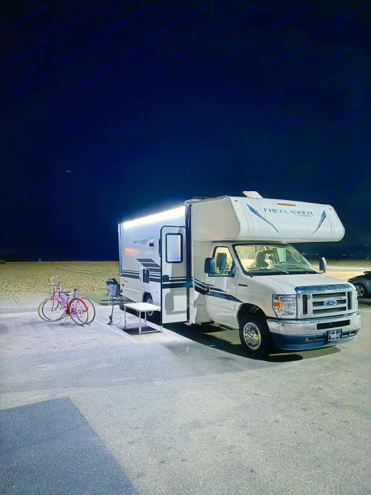 Illuminated RV rental parked at night by a beach with bicycles & an outdoor setup. A dog watches from inside.. Forest River Coachmen Freelander 2021