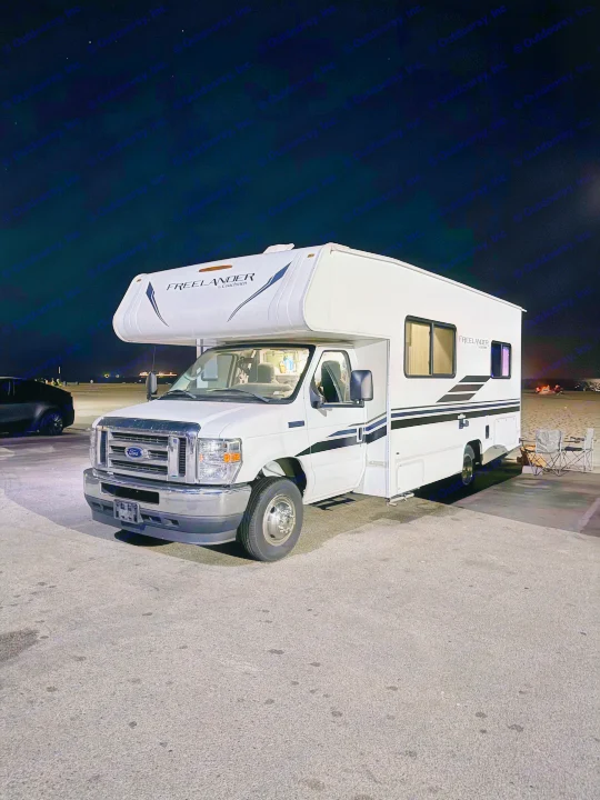 A white Coachmen Freelander Class C rv rental parked at night near a beach, with a distant campfire visible.. Forest River Coachmen Freelander 2021