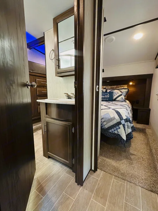 Step into this RV rental: a master bedroom with cozy blue/white bedding, visible from the adjacent bathroom vanity area and sink.. Forest River Cherokee Arctic Wolf 2019