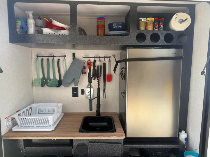 Organized RV rental galley kitchen with a compact fridge, sink, hanging utensils, and upper shelf storage.. Forest River Nobo 10.7 2024