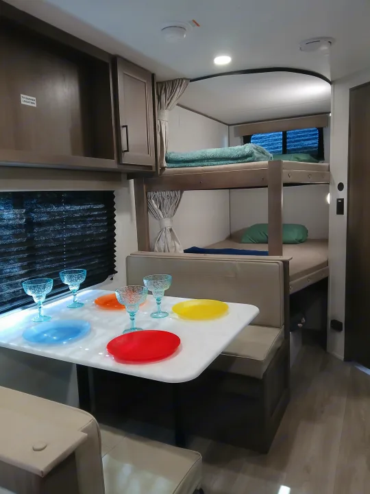 An RV rental's cozy interior shows a dinette with colorful plates and blue glasses, adjacent to practical bunk beds.. Grand Dessing Transend 2025