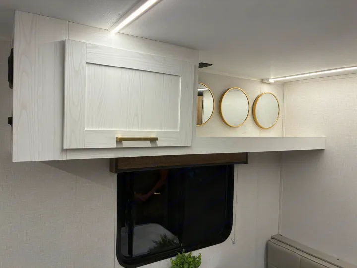Stylish RV rental interior with light wood-grain overhead cabinets, gold handles, circular mirrors, and LED lighting above a window.. Heartland RVs Eddie Bauer Edition 17RB-C 2025