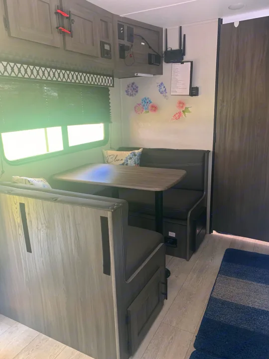 Inside an RV rental: a comfortable dinette with grey seating, wood table, overhead storage, and wall-mounted electronics.. Forest River Shasta 2021