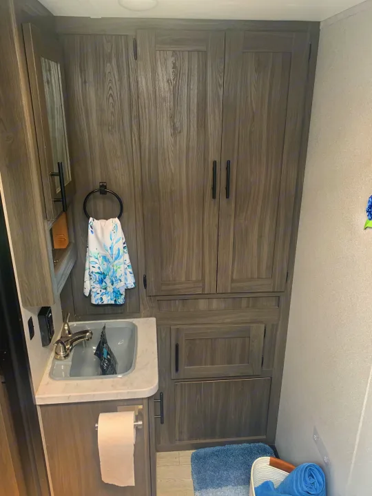 RV rental bathroom with a sink, mirror, wood-look cabinets, TP holder, and blue towels.. Forest River Shasta 2021