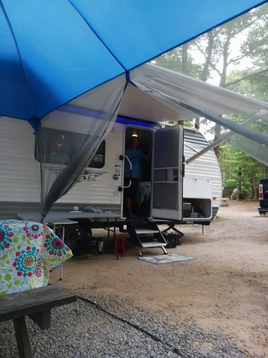 Man in an RV rental with blue awning extended at a dirt and gravel campsite, featuring a vibrant picnic table.. Forest River Shasta 2021