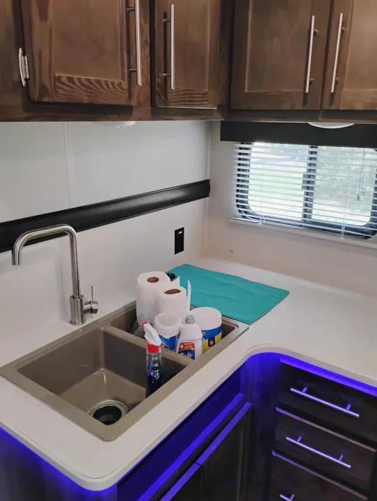 Kitchen area of an RV rental, showcasing a sink, wood cabinets, and vibrant blue under-counter lighting.. Crossroads RV Volante 2019