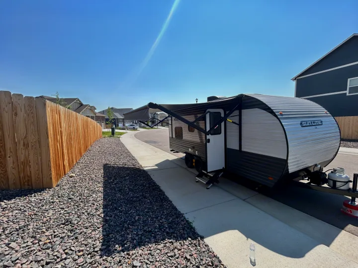 RV rental travel trailer with extended awning, parked on a driveway next to a wooden fence on a bright, sunny day.. Sunset Park & Rv Inc. 21bt 2026