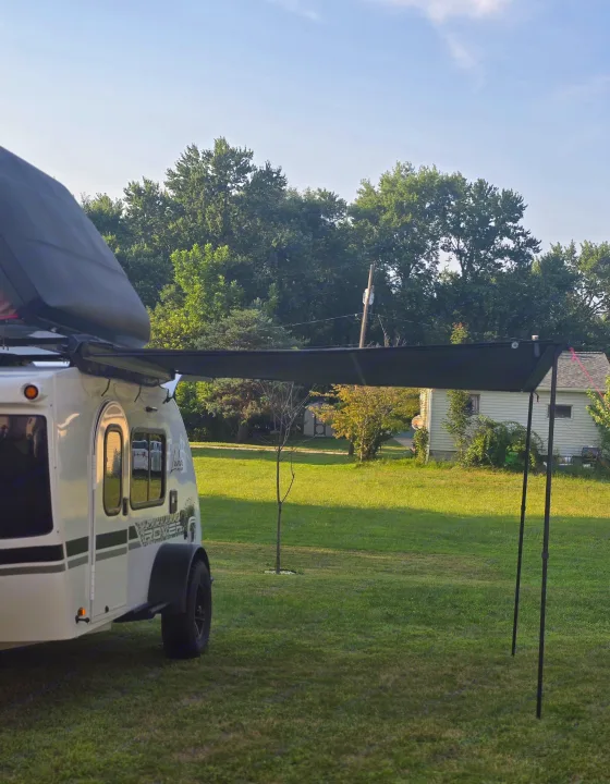 White teardrop RV rental with an extended black awning parked on a green lawn, ready for relaxing outdoor travel.. InTech RV Luna 2023