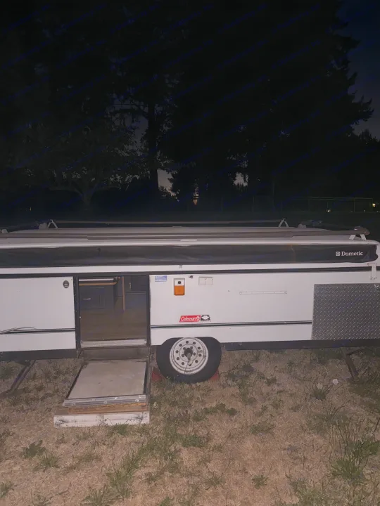A white pop-up RV rental with an open door showing its interior, parked on dry grass at night, silhouetted by dark trees.. Coleman Grand Tour 2002