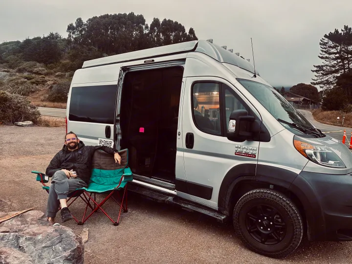 Man relaxes in a chair beside a silver Ram ProMaster RV rental on a gravel road, surrounded by trees.. Thor Motor Coach 18T 2022