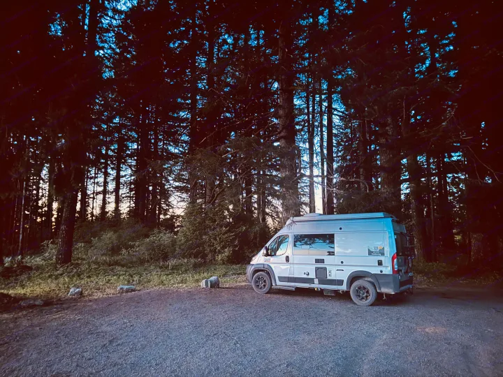 An RV rental camper van parked on a gravel path in a tall forest at sunset, perfect for outdoor travel.. Thor Motor Coach 18T 2022