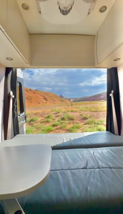RV rental interior view: a cozy bed & table frame a scenic desert landscape with hills and road.. Pleasure-Way Pleasure-Way Class B 2023