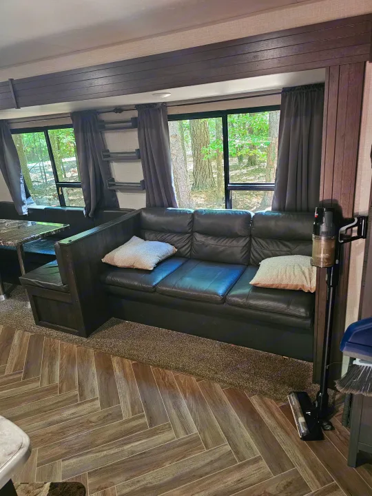 Cozy RV rental interior with dark leather sofa, dining area, and windows showing a wooded view, perfect for relaxation.. KZ Sportsman 2021