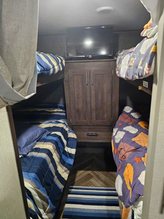 Inside an RV rental, showing compact bunk beds with striped and s'mores bedding, central TV, and cabinet storage.. KZ Sportsman 2021