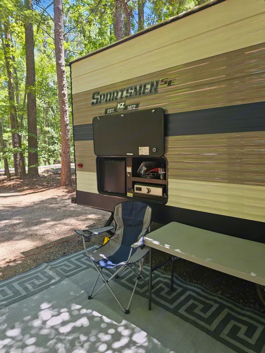 RV rental campsite setup: outdoor kitchen, chair, and table on a patterned rug amidst sunlit woods.. KZ Sportsman 2021