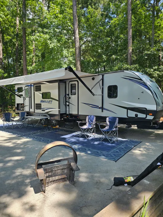 Enjoy a serene rv rental experience! A white travel trailer is set up at a shaded campsite with an awning, chairs, and fire pit.. Keystone RV Bullet Ultra Lite 2019