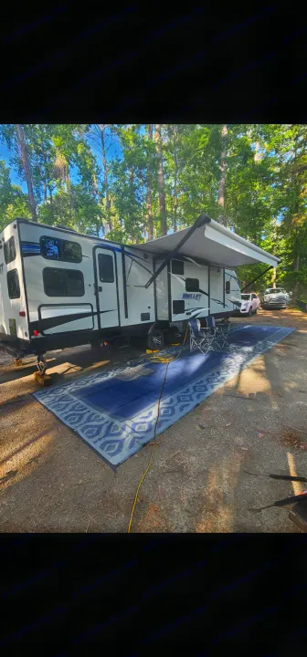 RV rental at a wooded campsite: a white travel trailer with awning, blue outdoor rug, and chairs set up for relaxation.. Keystone RV Bullet Ultra Lite 2019