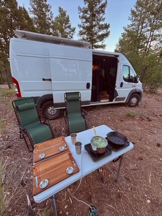 RV rental parked in a forest clearing with outdoor cooking setup: pot, pan, cutting boards, and chairs.. RAM Promaster 2500 2018