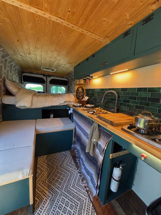 Cozy converted van interior for RV rental: bed, kitchen with green tiles, wood ceiling, and patterned rug.. RAM Promaster 2500 2018